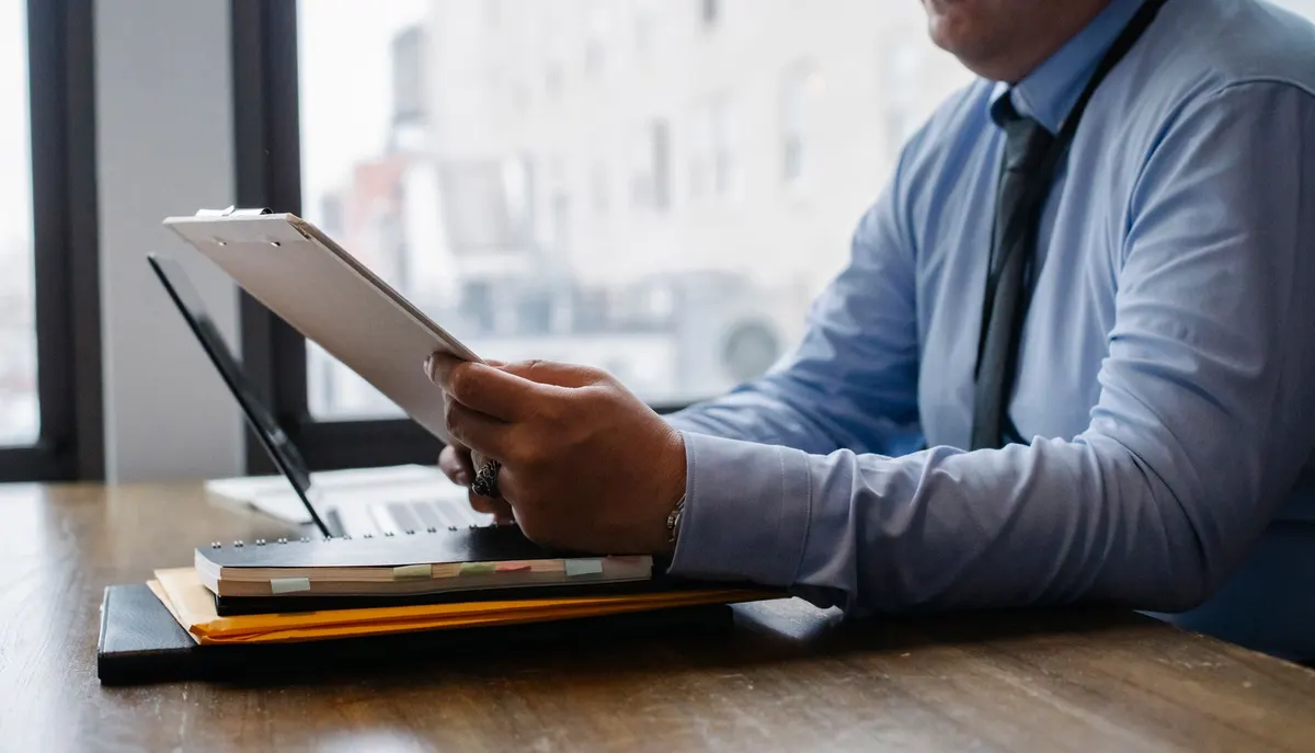 A man in a blue shirt reading from a clipboard at a desk with folders and a laptop