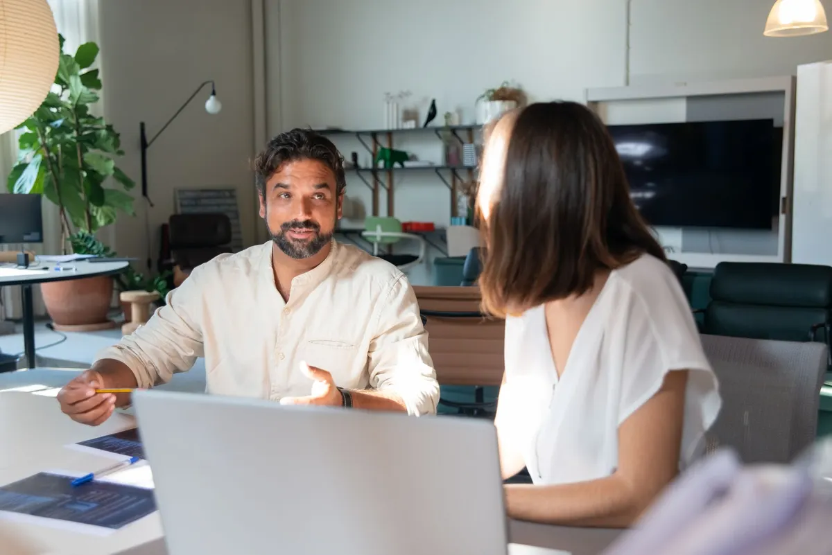 Two colleagues talking across a desk with a laptop open between them