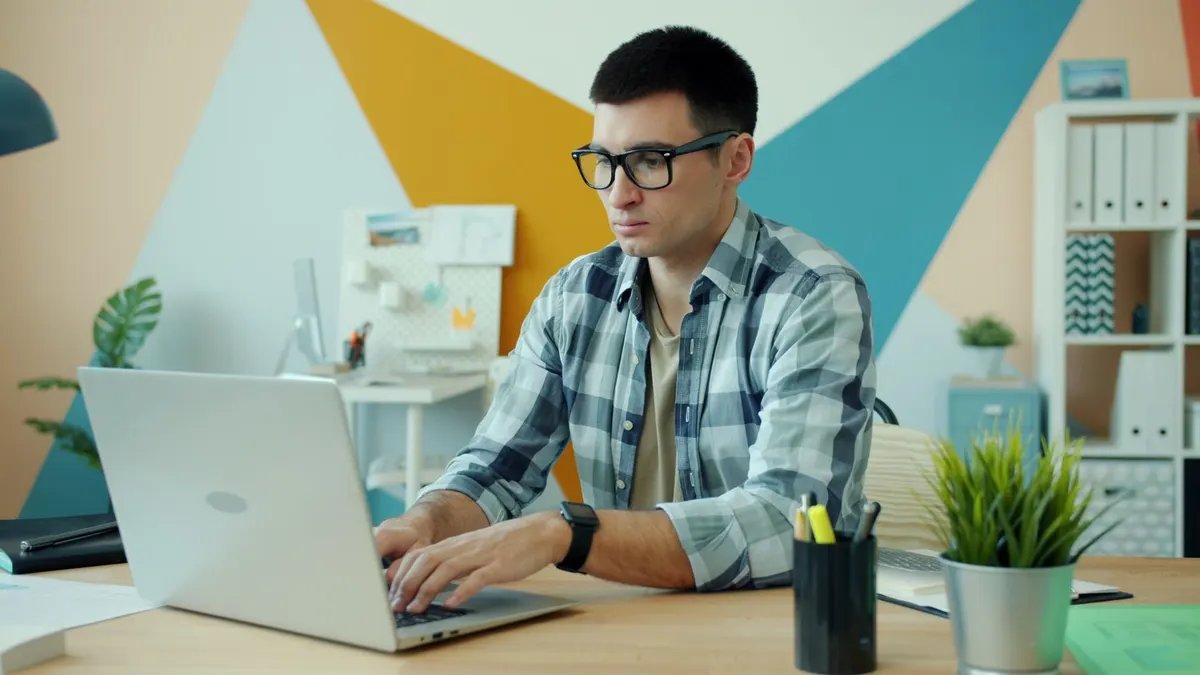A man in a plaid shirt working at a laptop on a desk with a small plant and pens