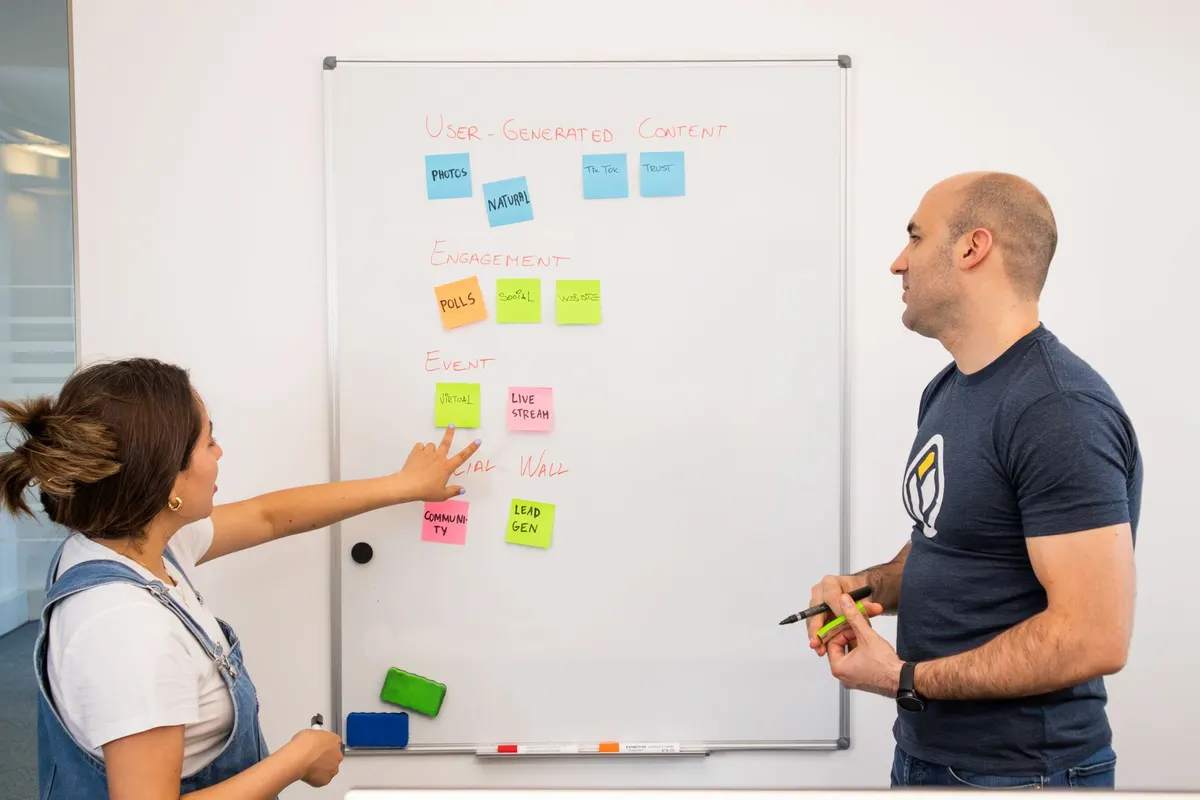 A woman and a man at a whiteboard covered in sticky notes, pointing at the columns