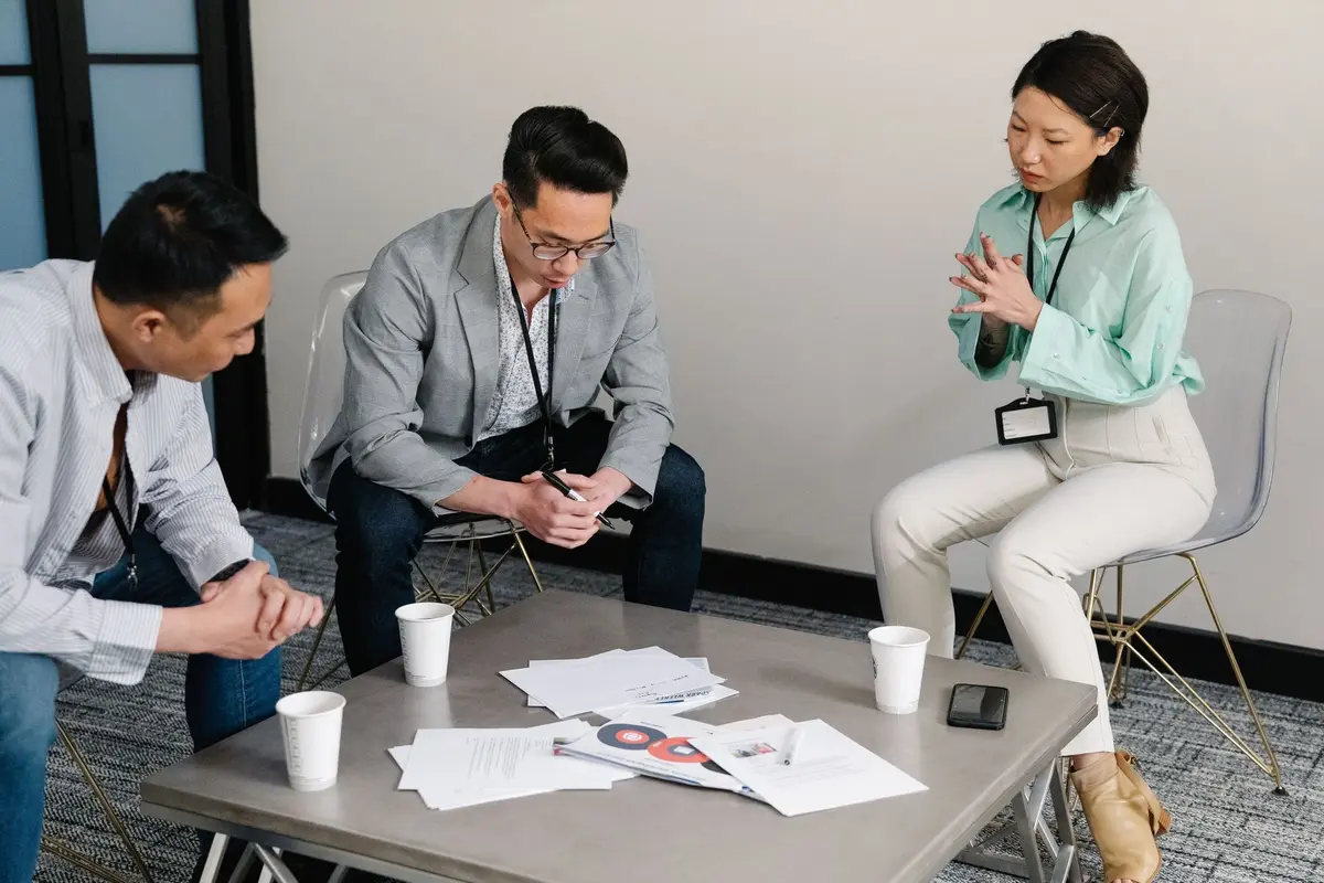 A group of colleagues in a meeting room discuss papers on a table during a business discussion.