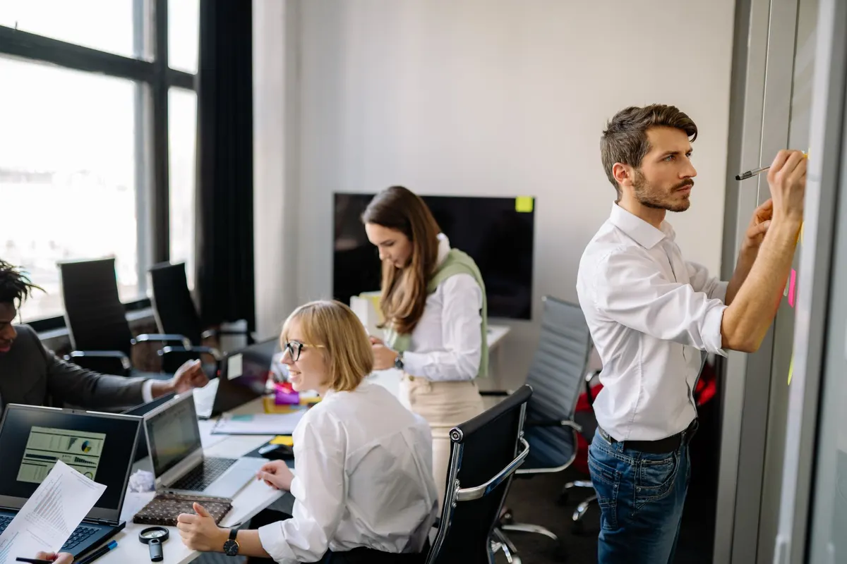 A man at a wall with sticky notes while colleagues work at laptops behind him