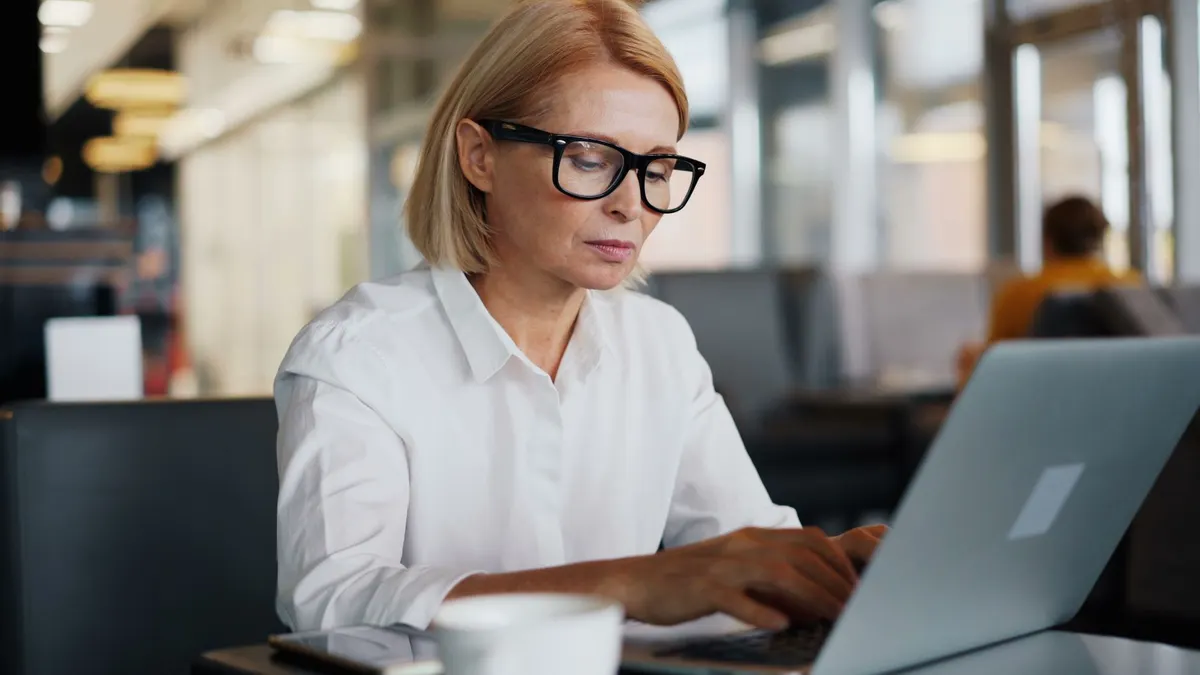 A woman with glasses typing on a laptop at a desk in a bright office