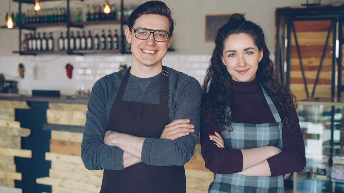 Two cafe owners in aprons standing side by side with arms crossed behind a counter