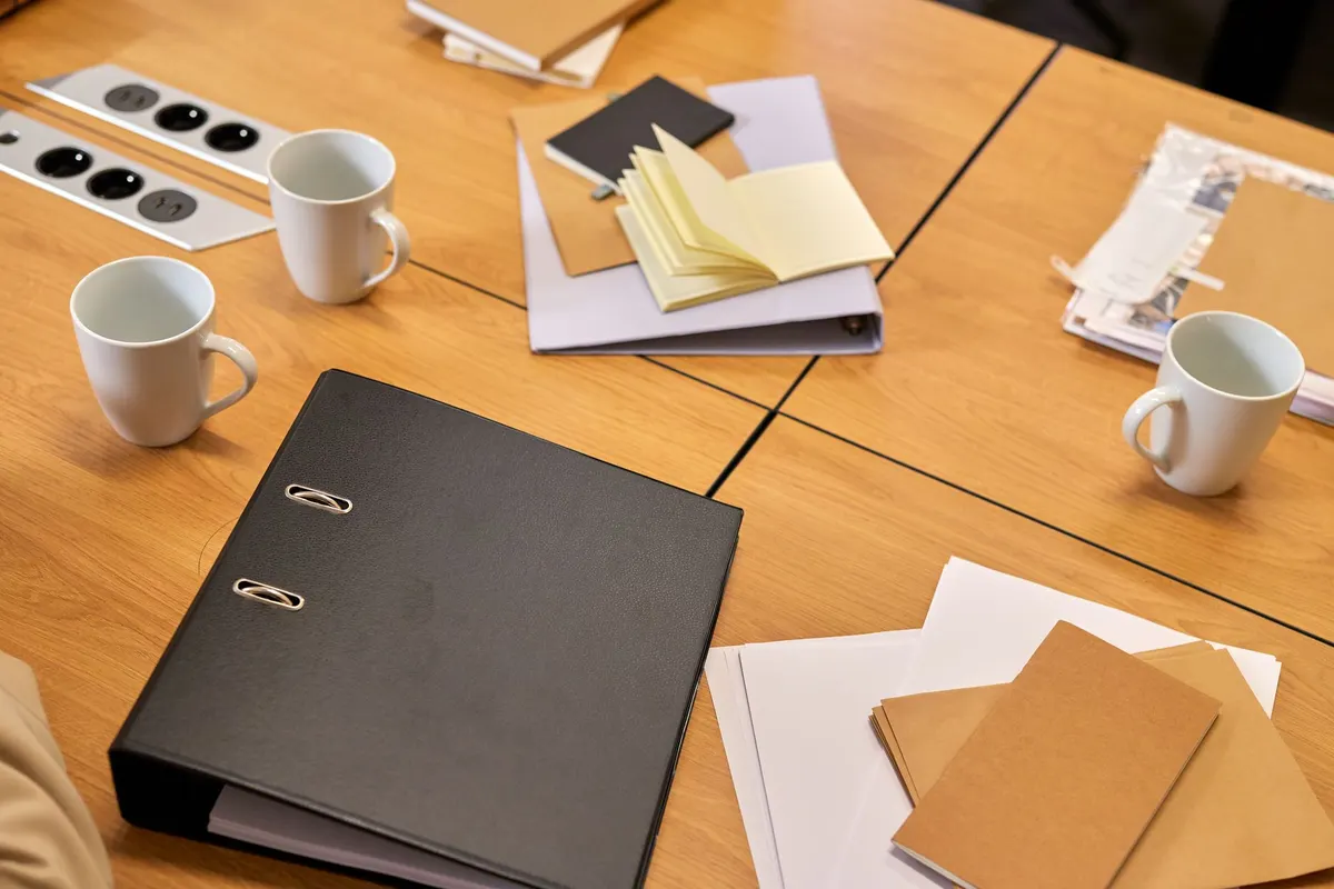 A black binder, notebooks, and coffee mugs on a wooden desk
