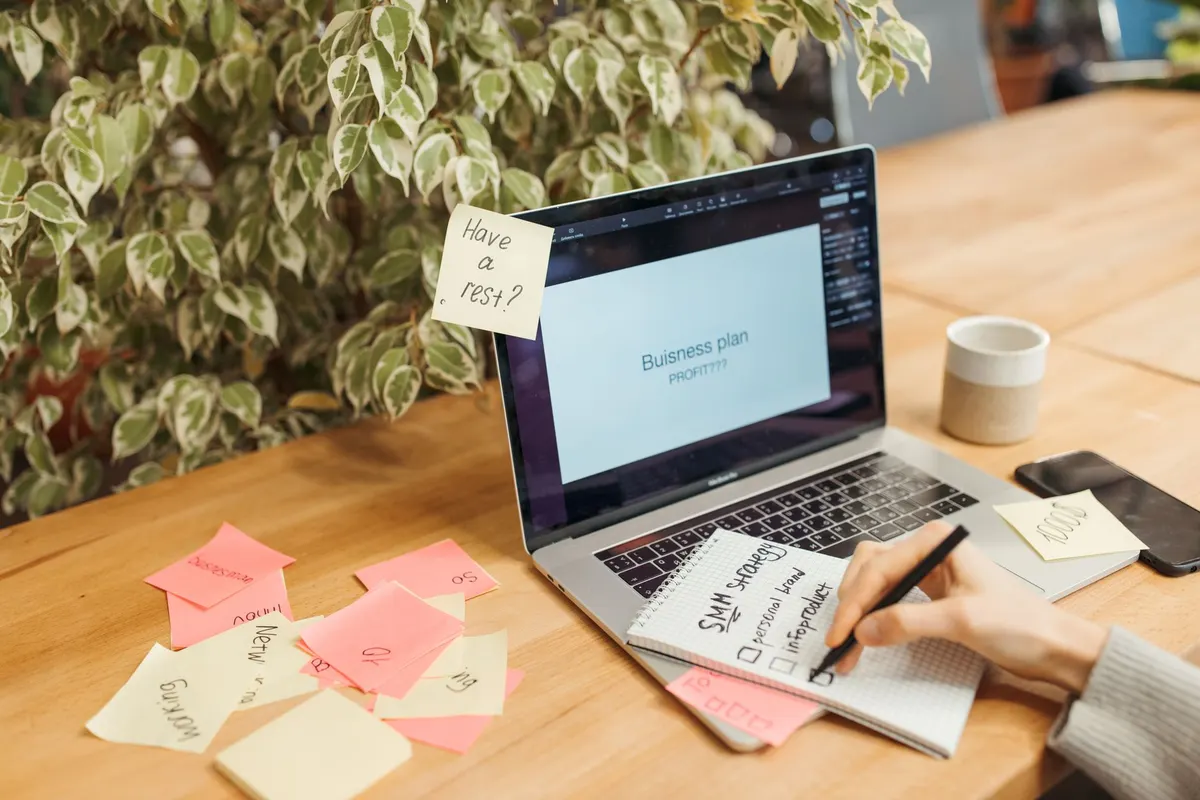 A laptop, a notebook and sticky notes on a wooden desk while a hand writes a note