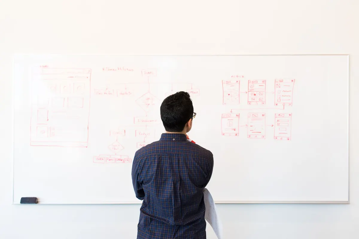 A man seen from behind standing at a large whiteboard, thinking over a plan