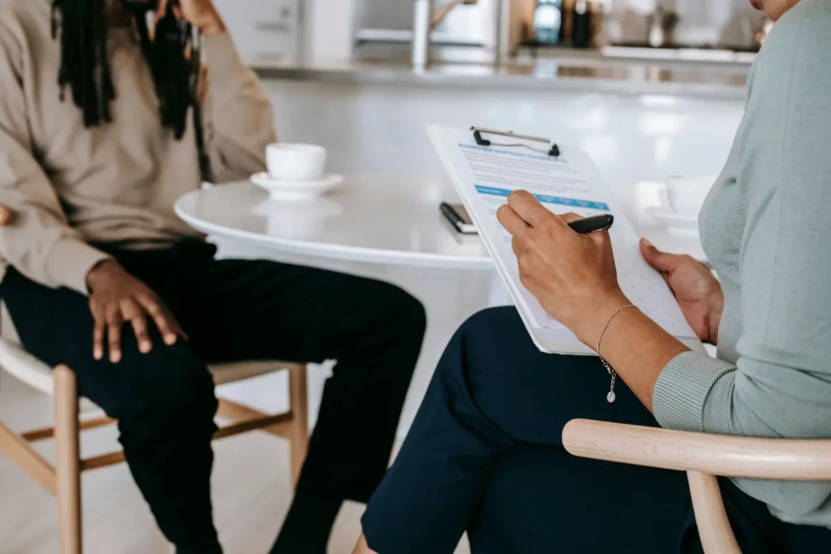 Crop unrecognizable female psychotherapist or interviewer asking questions and taking notes in clipboard during interview with black male.