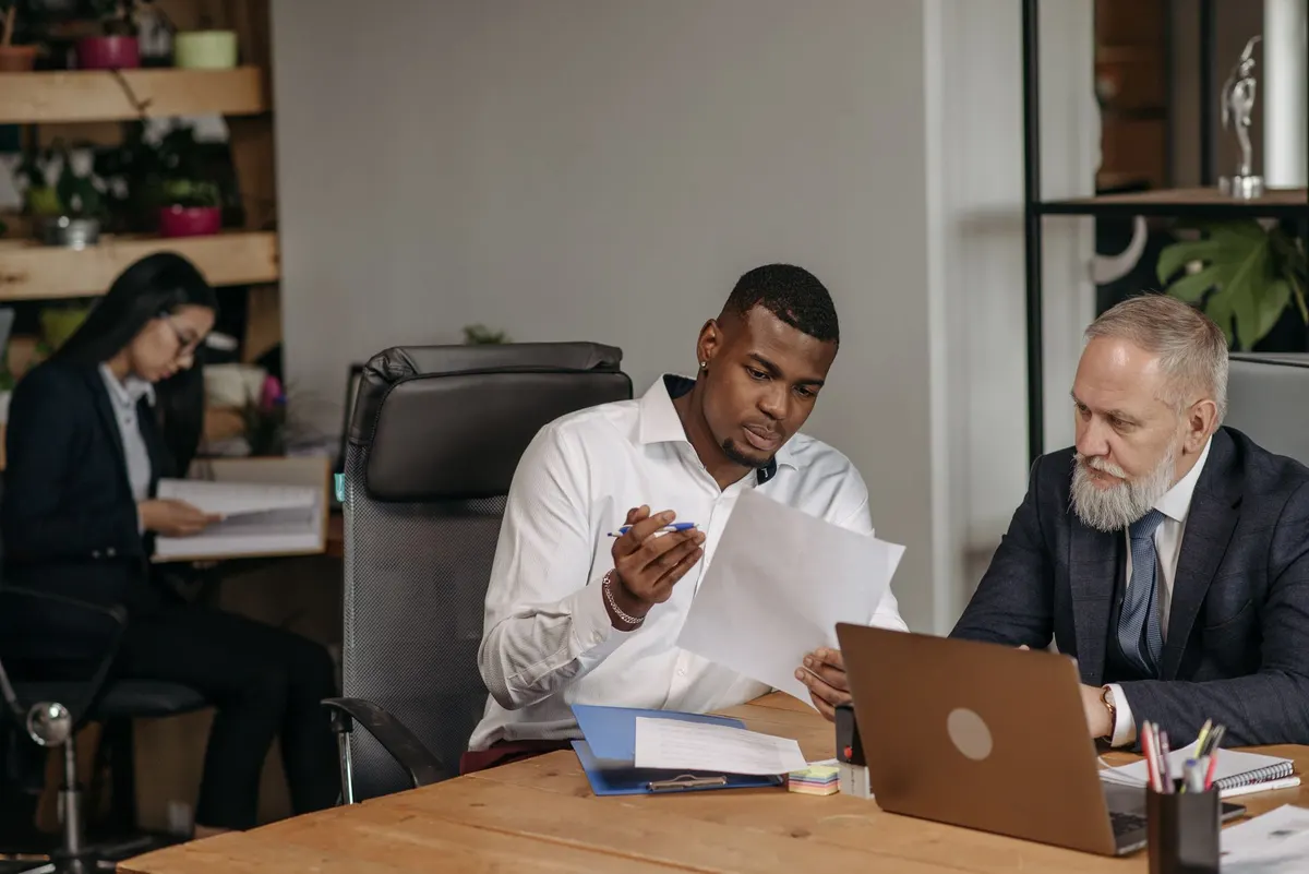 Two men at a desk with a laptop, one reading a printed document