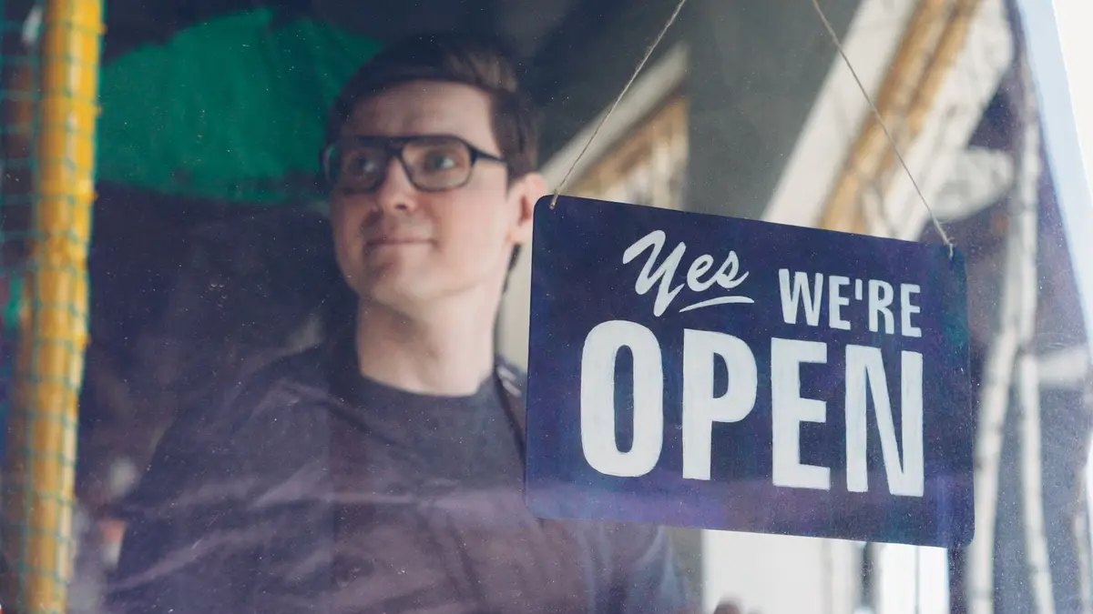 Smiling adult man behind a glass with a 'Yes, We're Open' sign.