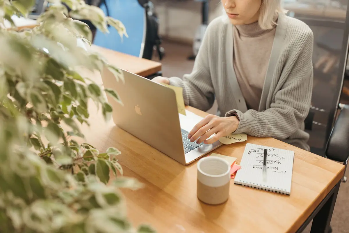A woman working on a laptop at a wooden desk in a bright, modern office setting.