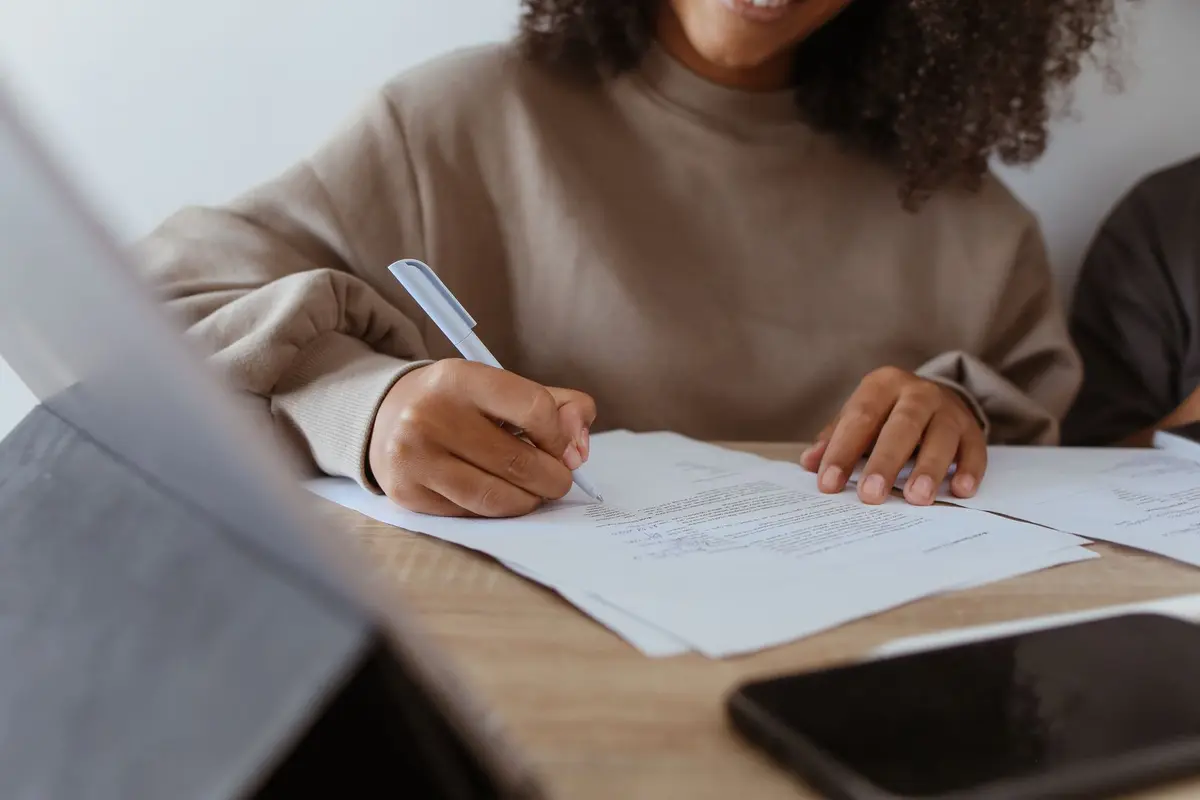 Close-up of a woman's hands writing on documents with a pen indoors.