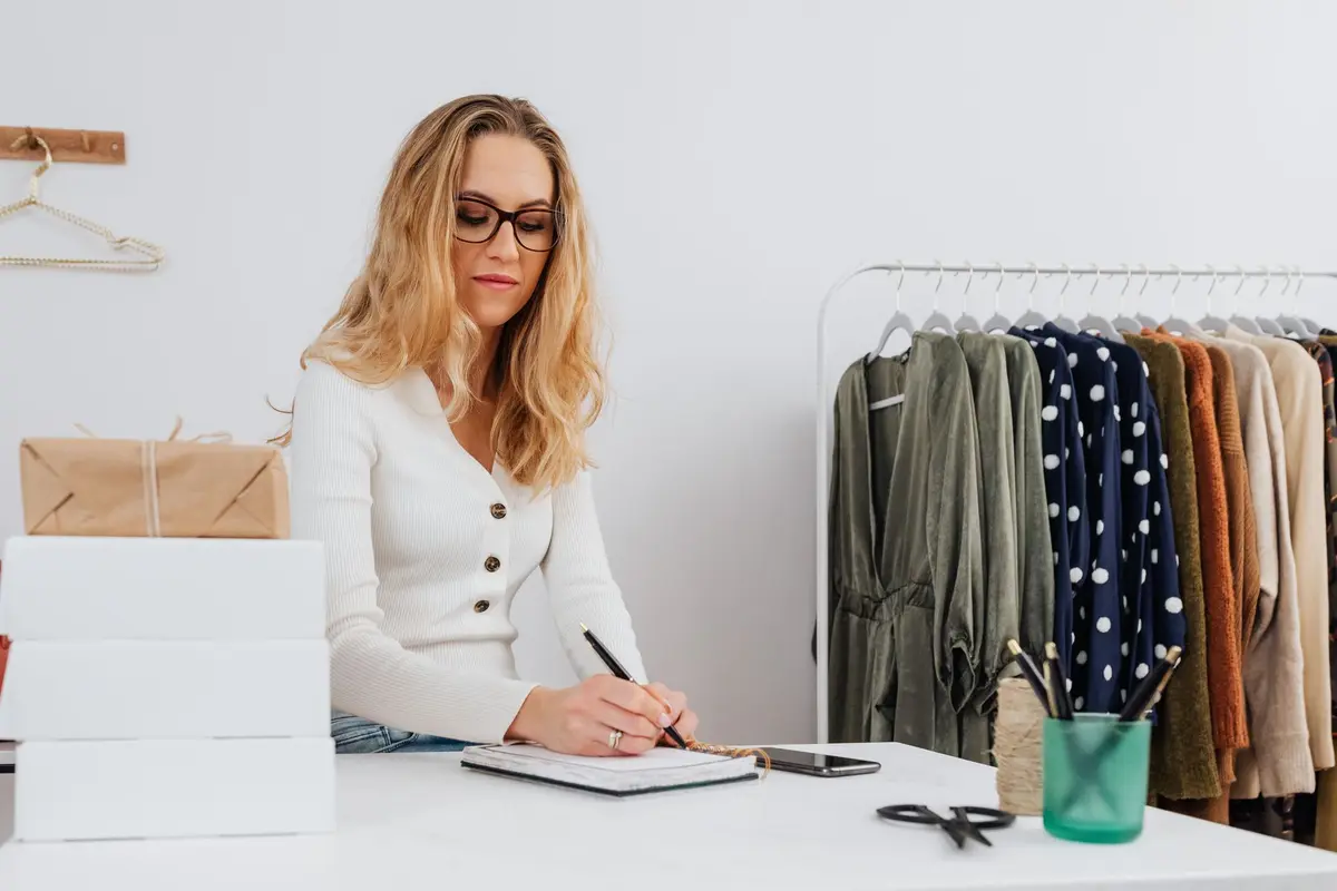 Caucasian woman in white sweater writing at desk with clothes rack.