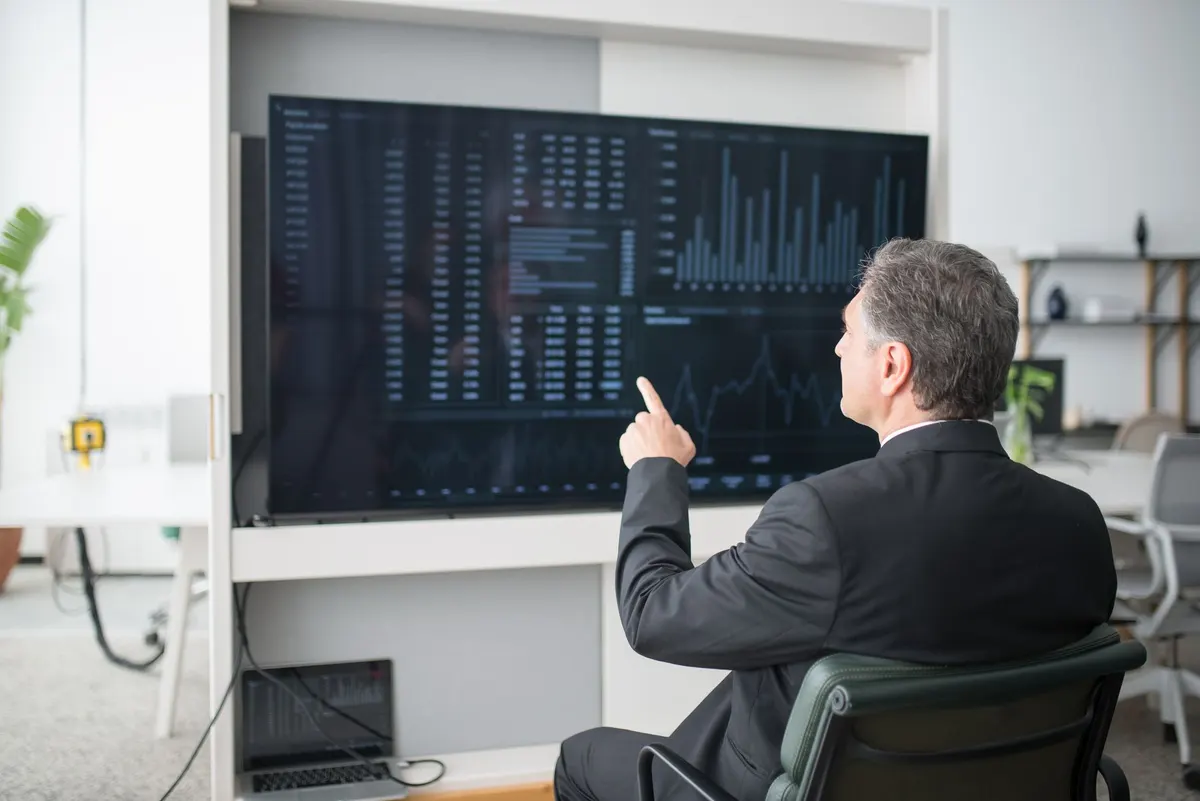 A man seated in a meeting room pointing at a wall screen of charts