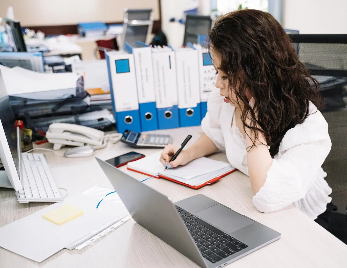 A woman writing in a notebook at a desk with binders, a laptop and a phone