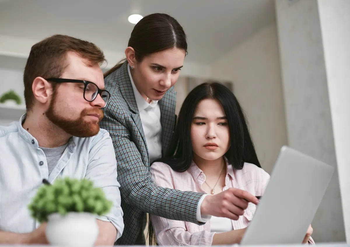 Three colleagues leaning in to look at the same laptop, one pointing at the screen