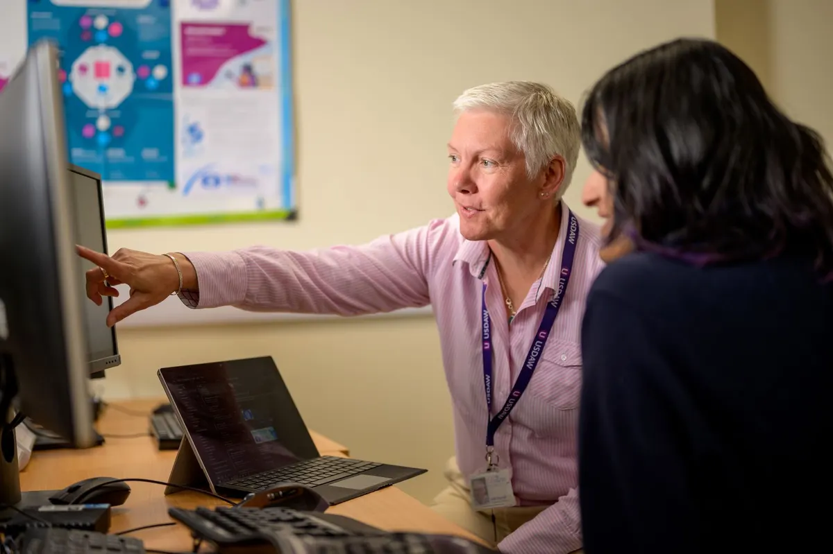 An older man with a lanyard pointing at a screen while explaining it to a colleague