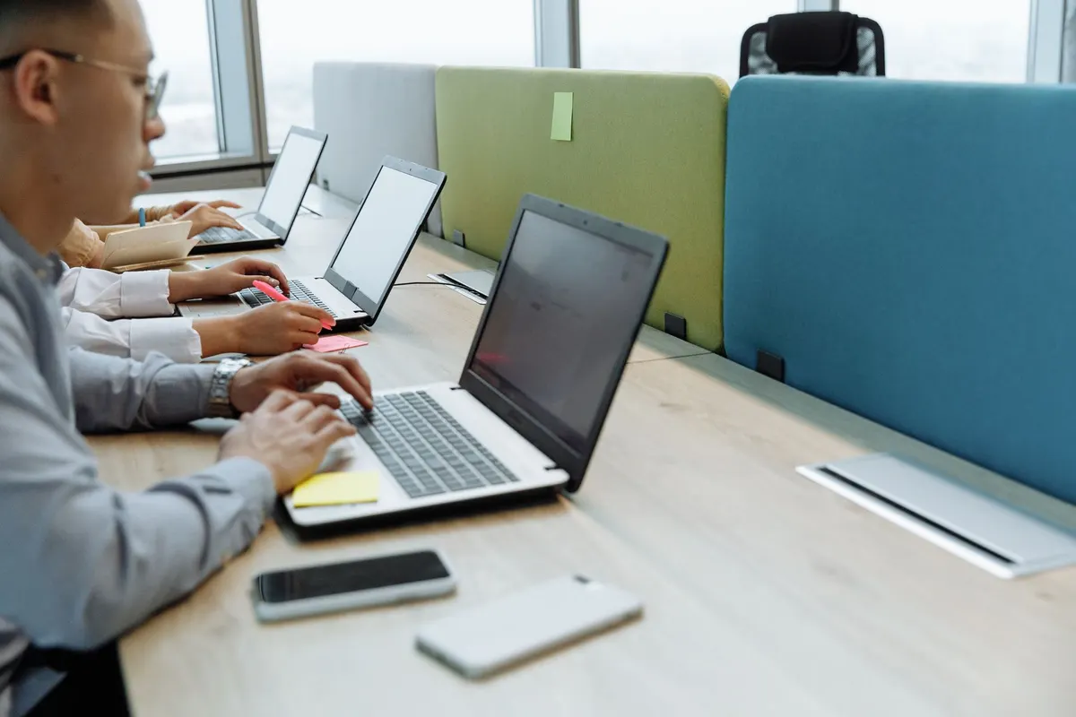 A row of people typing at laptops along a shared desk with partitions
