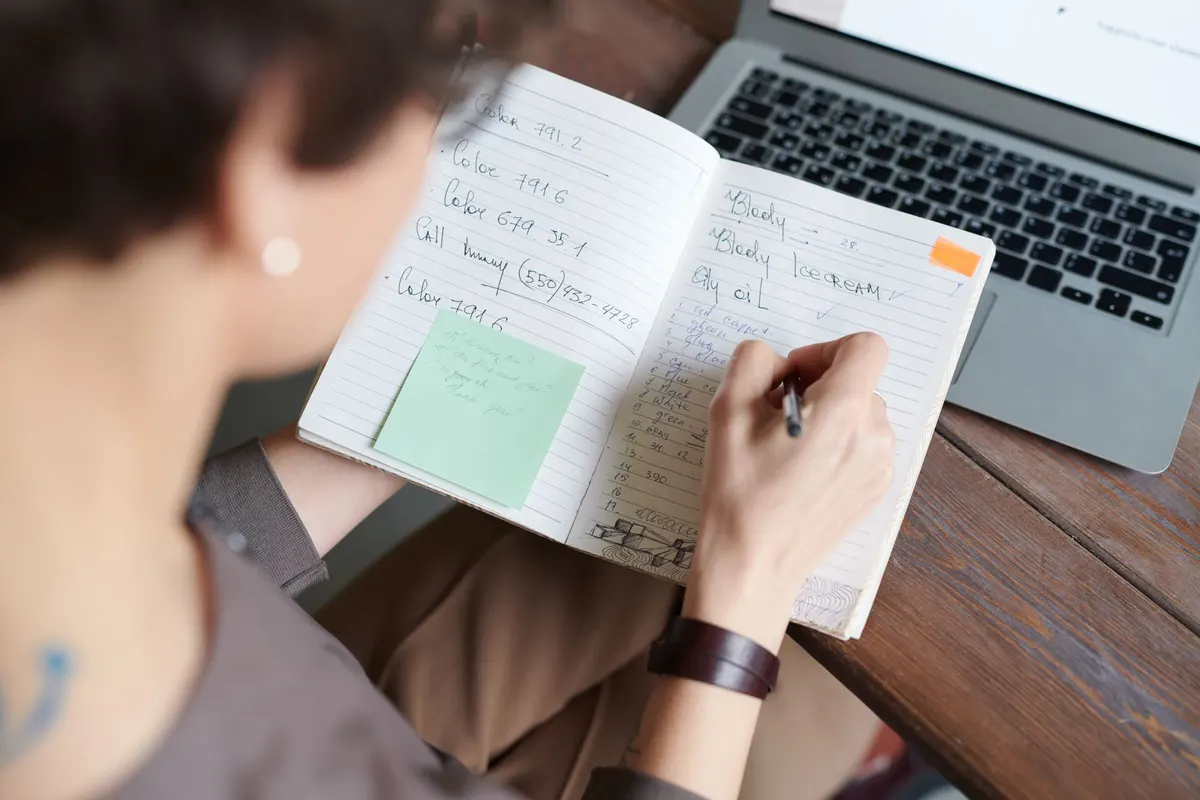 A person writing in a notebook next to an open laptop, with a sticky note on the page